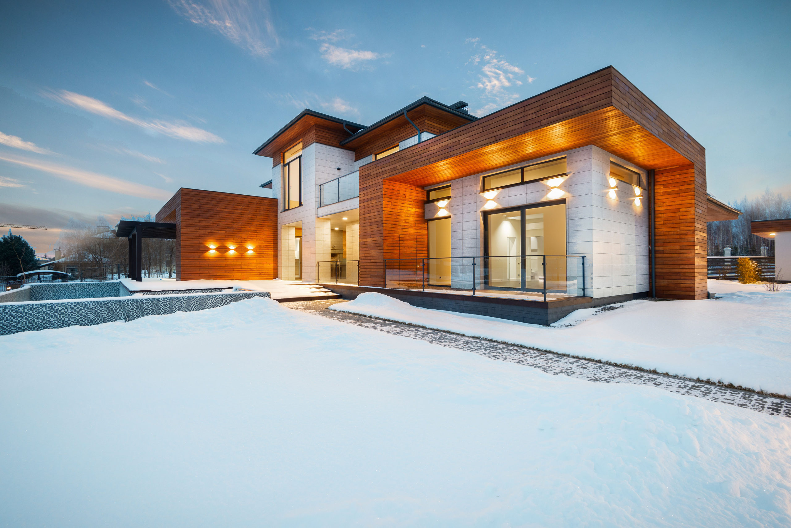 A modern stone and cedar house lit from within at dusk, deep snow across the ground