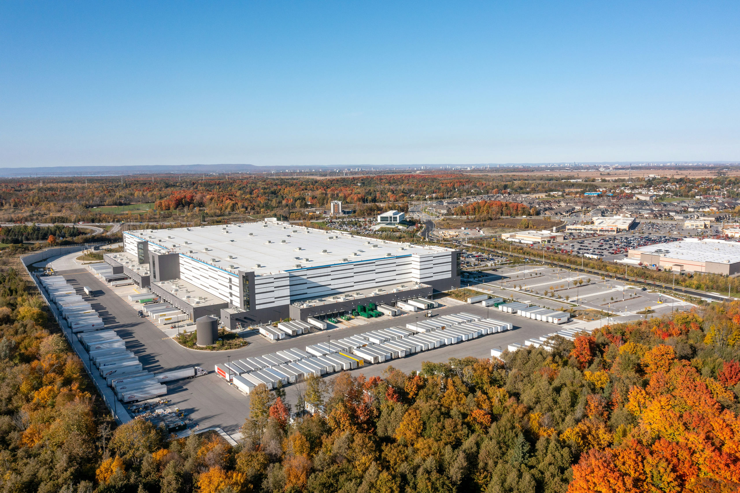 An aerial view of a large distribution building, its loading docks lined with trailers, parking beyond it and autumn woodland all around